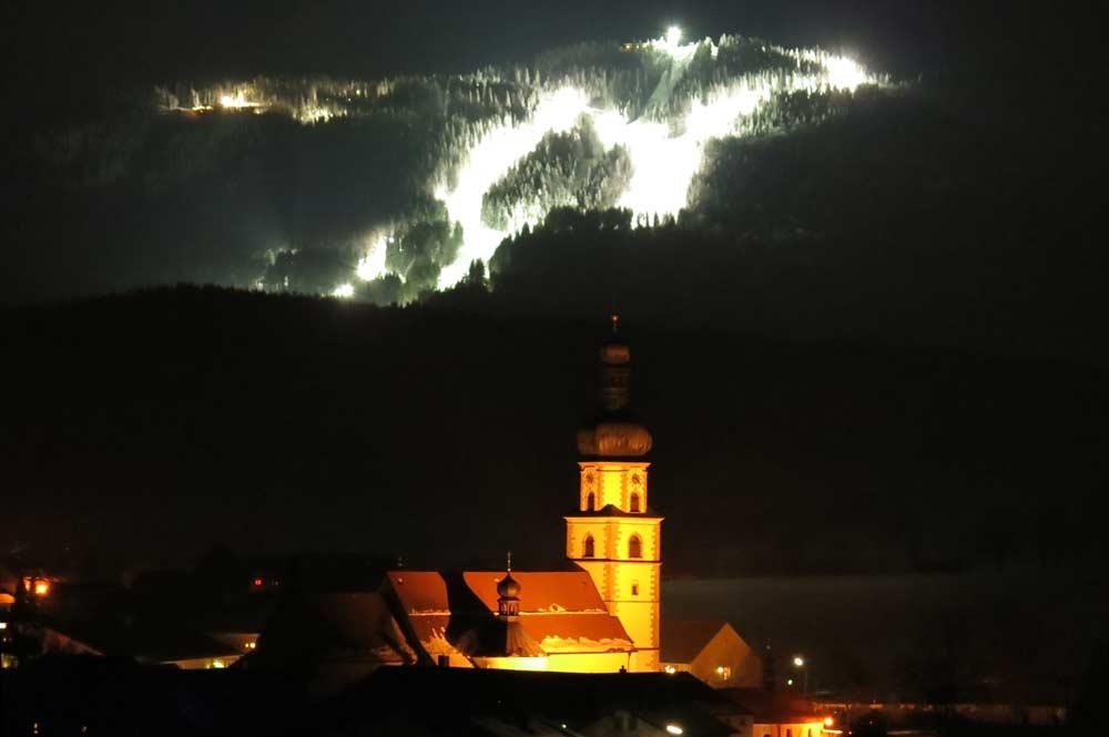 Beleuchtete Wallfahrtskirche mit der beleuchteten Piste im Hintergrund